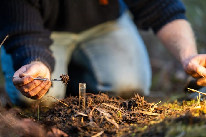 A person can be seen inspecting the soil with a test tube planted into the ground and adding soil into that test tube