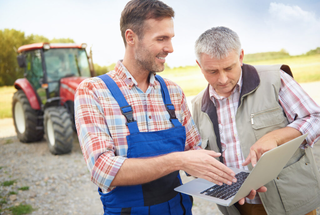 Two men can be seen discussing something and one of them is holding a chart, there's a tractor can be seen seen in the background as well